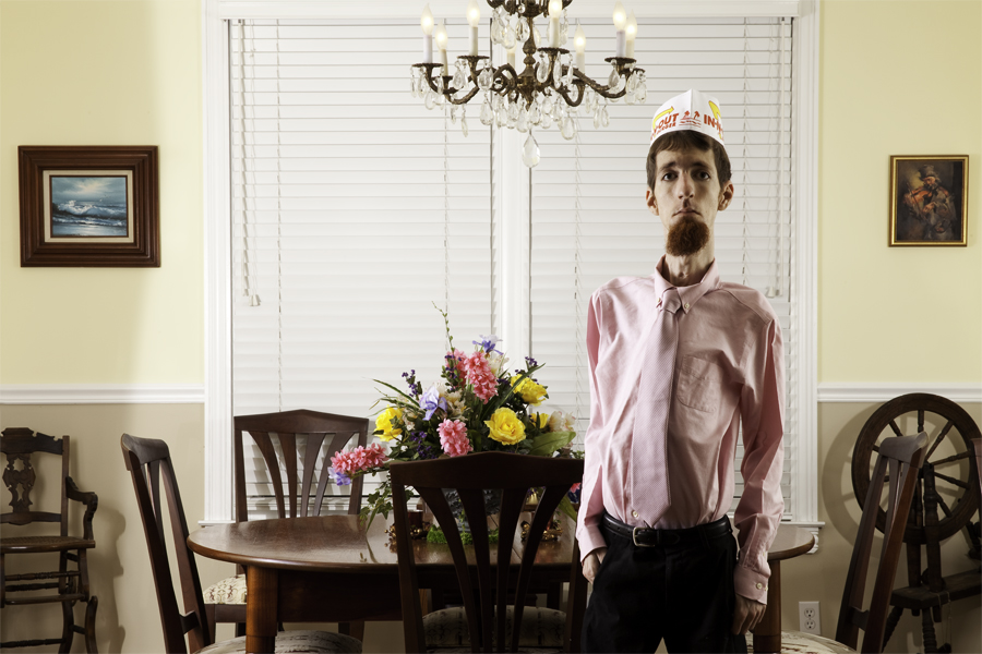 This Brian Charles Steel photo is a self-portrait of the artist in a dining room. He is standing in front of the wooden dining room table with his hands in his pockets.  The table has a vase of flowers on top of it. Above the table is a chandelier.  He is lit dramatically; both side so him are well lit, but his front is in shadow.  He is wearing black dress pants, pink dress shirt, a pink tie, and an In And Out cap.  His hair is short and brown. On the wall behind him on the right there is an ocean painting and a wooden high chair. 